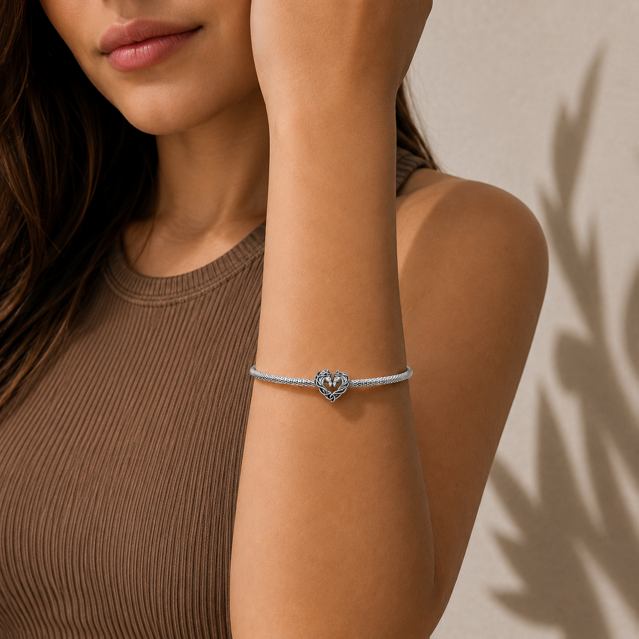 Close-up of a woman's arm wearing a silver bracelet with a horse heart charm on a beige background